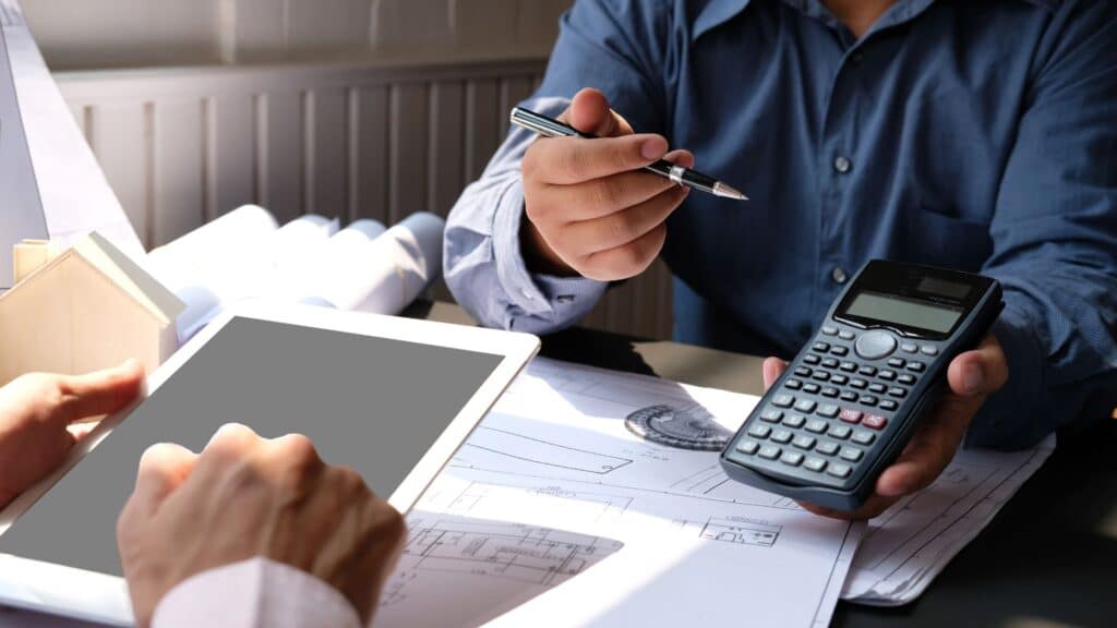 Two people working together at a desk, one holding a calculator and a pen, and the other using a tablet. Blueprints and documents are spread out on the desk.