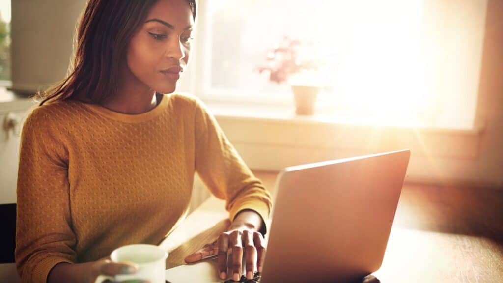 A woman in a yellow sweater sits at a table, focused on her laptop while holding a white mug, with sunlight streaming through a window behind her.