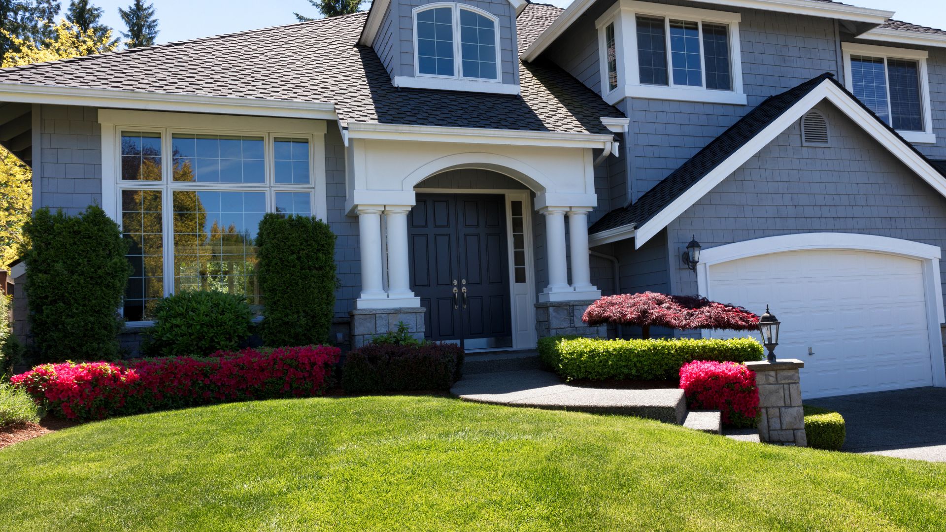 A modern two-story house with grey siding, a well-kept lawn, manicured shrubs, and a flower bed. The house has large windows and a black front door with white trim.